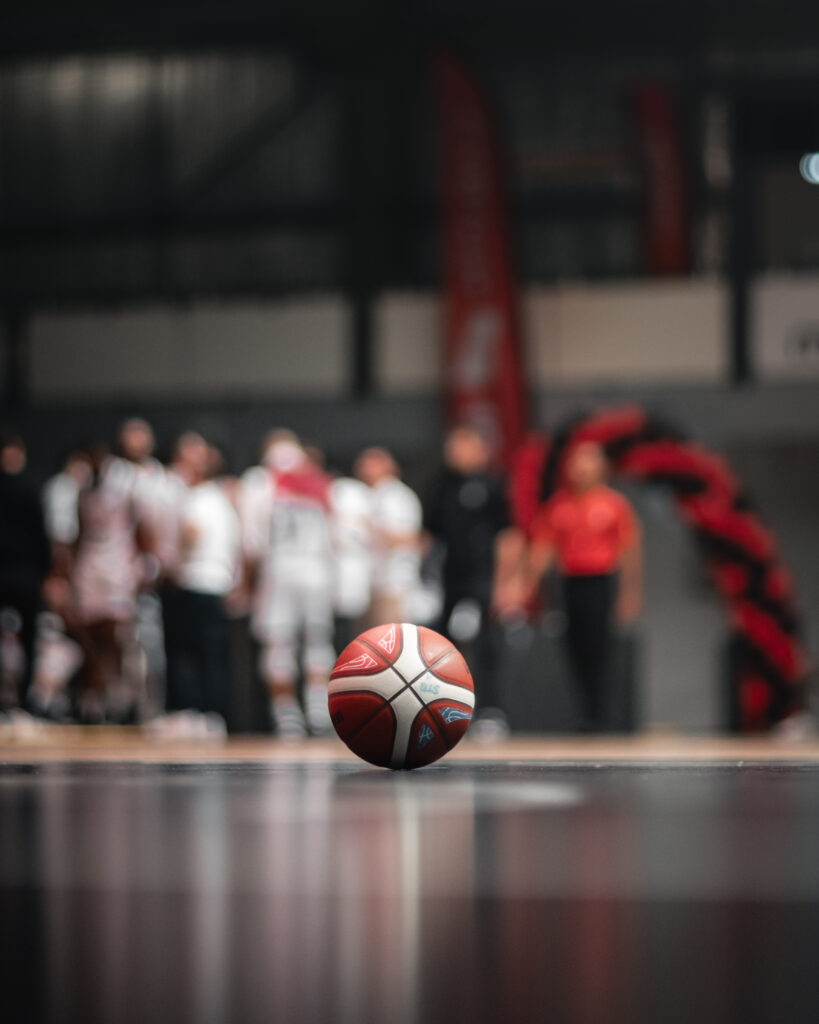 Photo d'un ballon de basket posé sur au millieu du terrain. En fond, un groupement de joueurs. La mise au point est faite sur le ballon le fond est flou